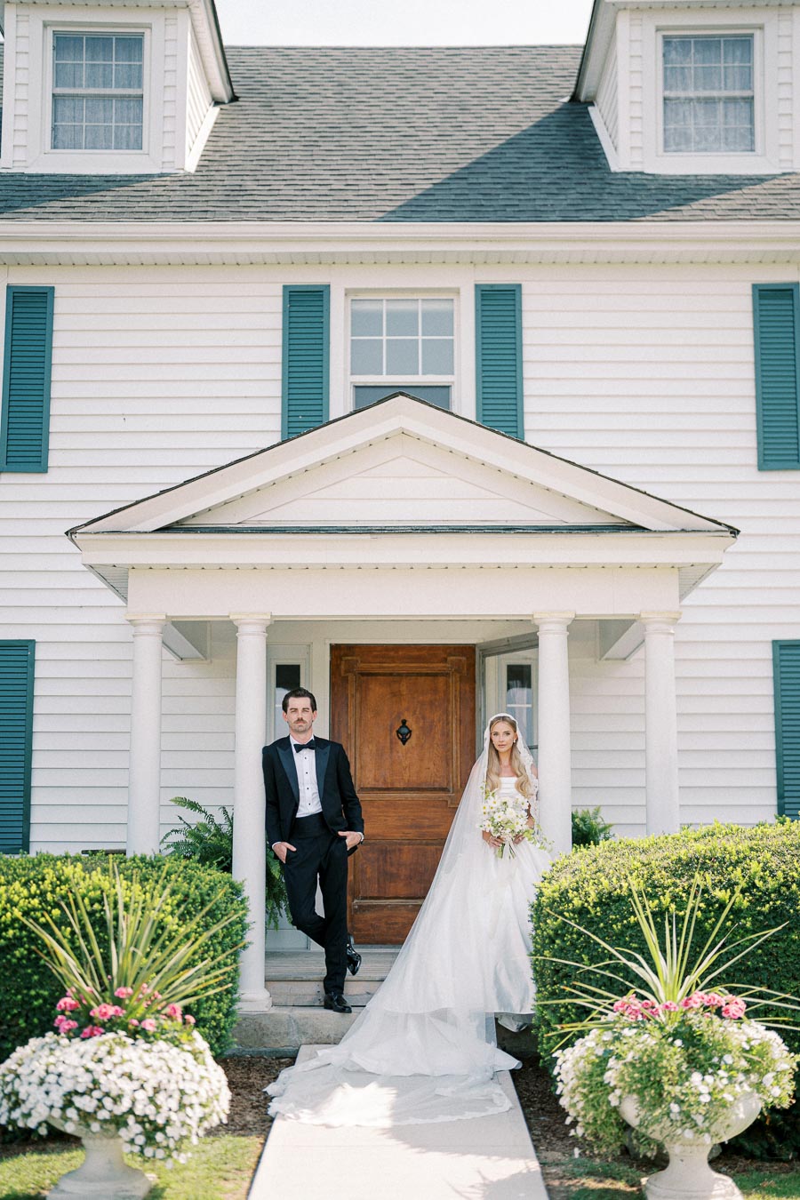 Elegant bride and groom posing in front of a charming white house with blue shutters, capturing timeless wedding memories.