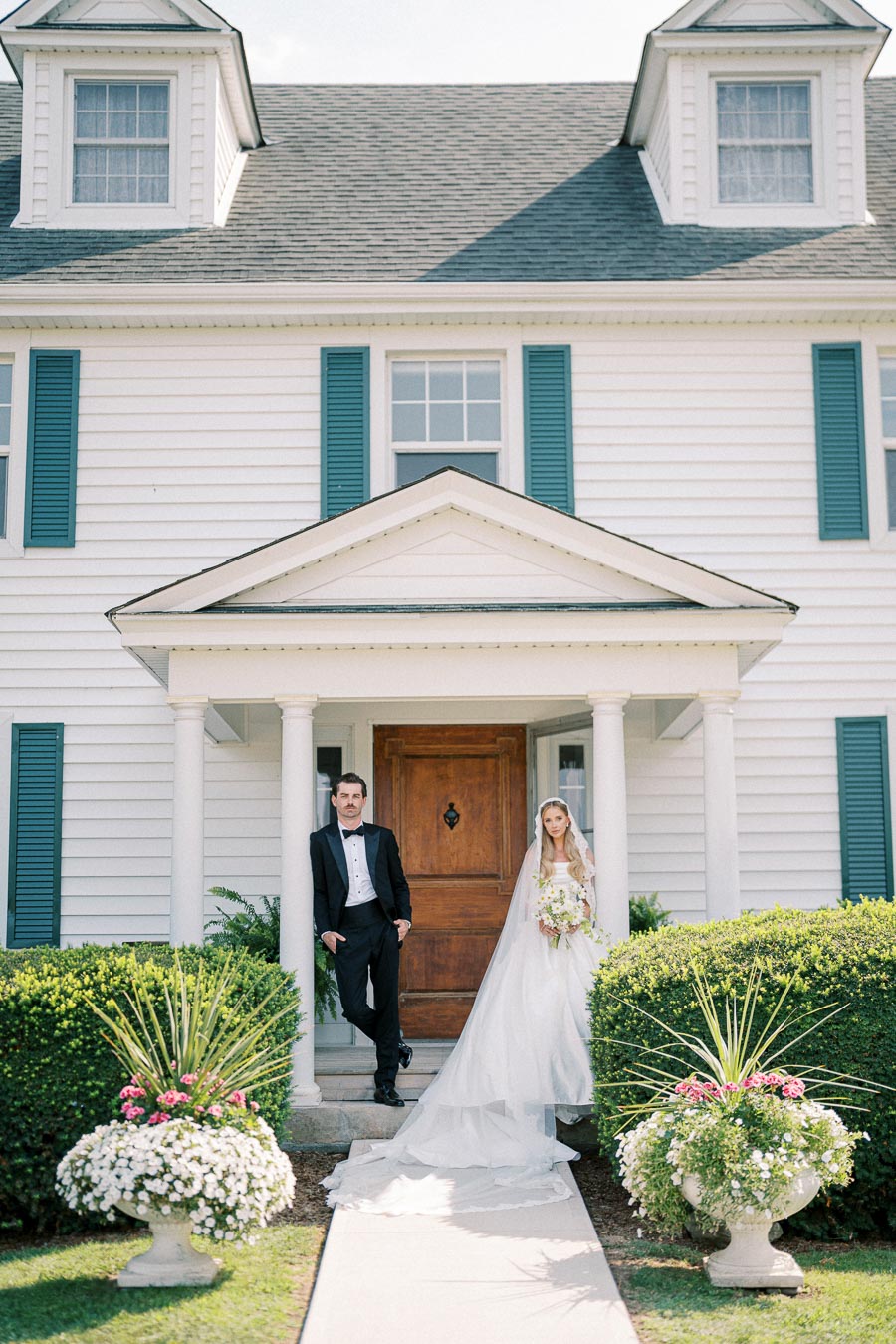 Bride and groom standing outside a classic white house with blue shutters, beautifully landscaped with greenery and flower arrangements, showcasing elegant wedding attire.