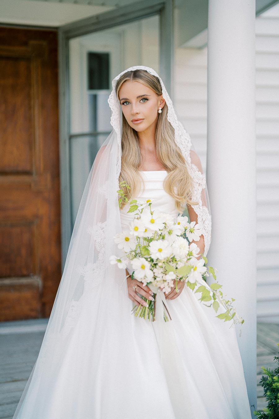 Bride in a white wedding gown holding a bouquet of white flowers, standing on a porch with a wooden door in the background.
