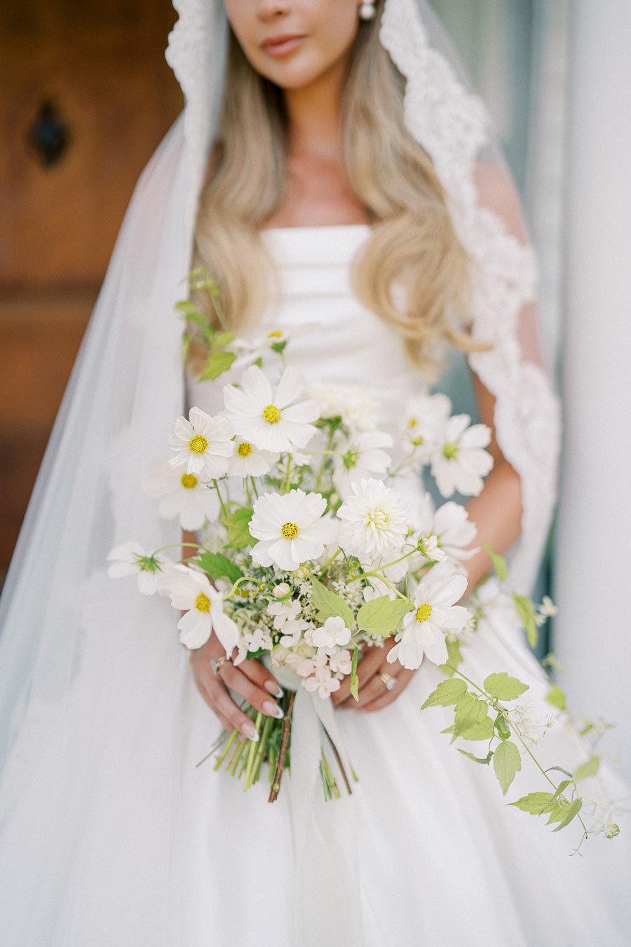 Bridal portrait with elegant white lace veil, long blonde hair, and a bouquet of white daisies and green foliage against a soft background.