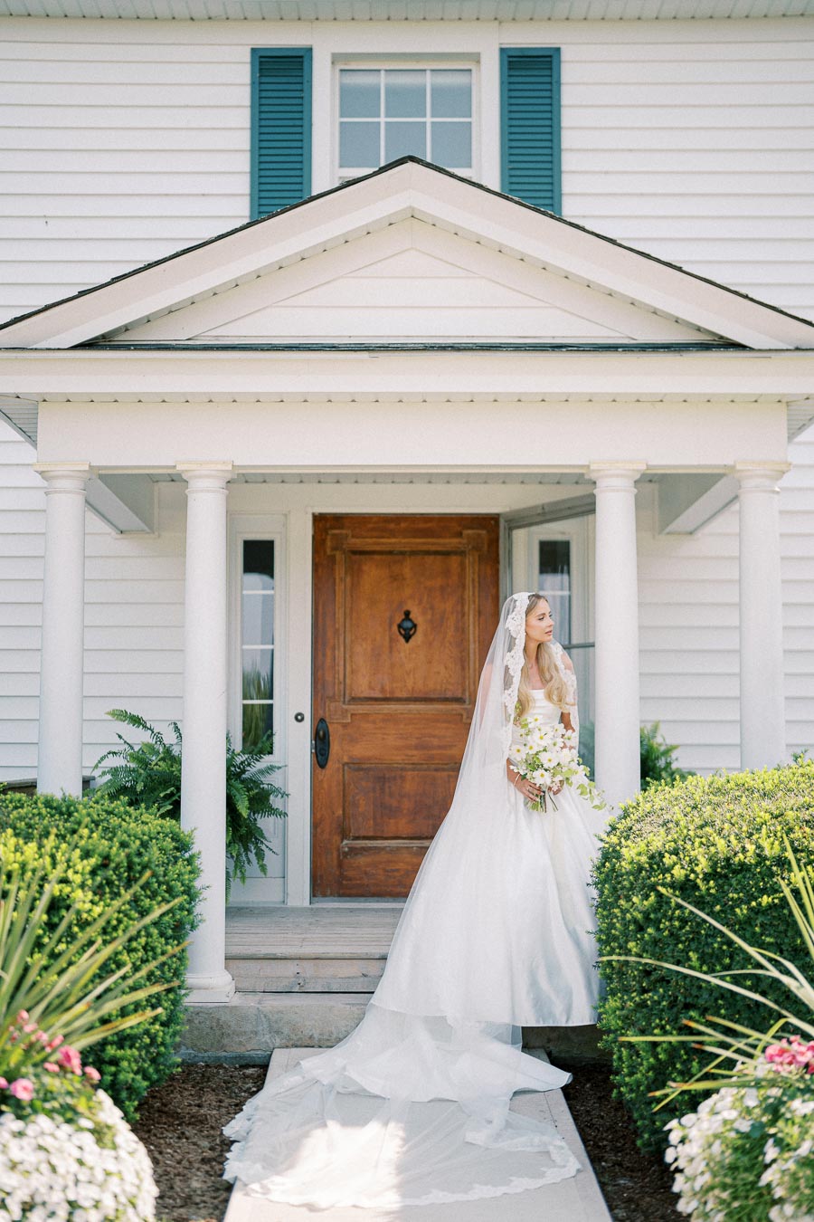 A bride in a flowing white gown and veil stands in front of a charming wooden door with columns, holding a bouquet of white flowers, surrounded by lush greenery and a neatly manicured garden.