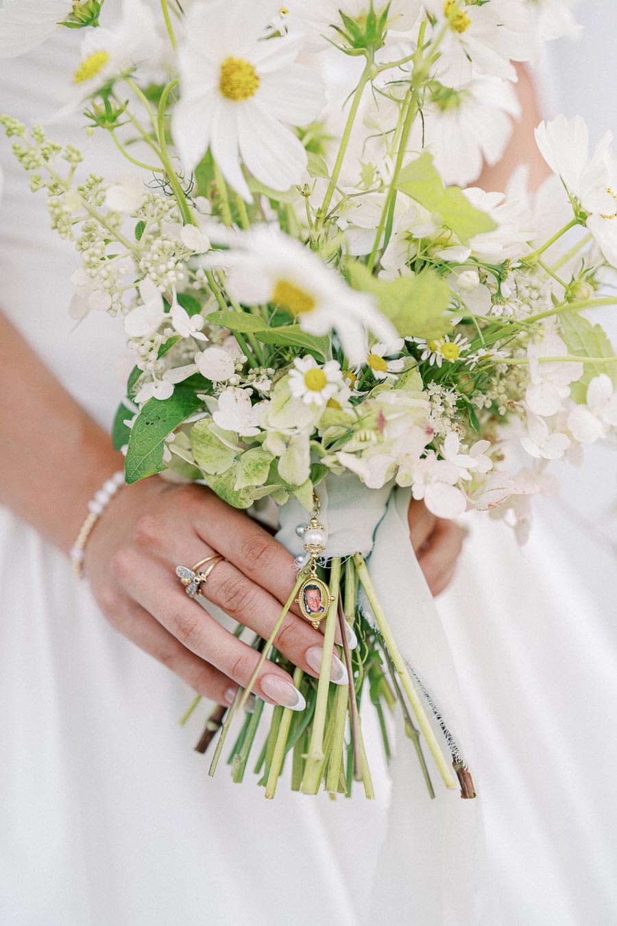 Bridal bouquet with white daisies and greenery held by a bride wearing a pearl bracelet and a ring, featuring a small photo pendant as a personal touch.