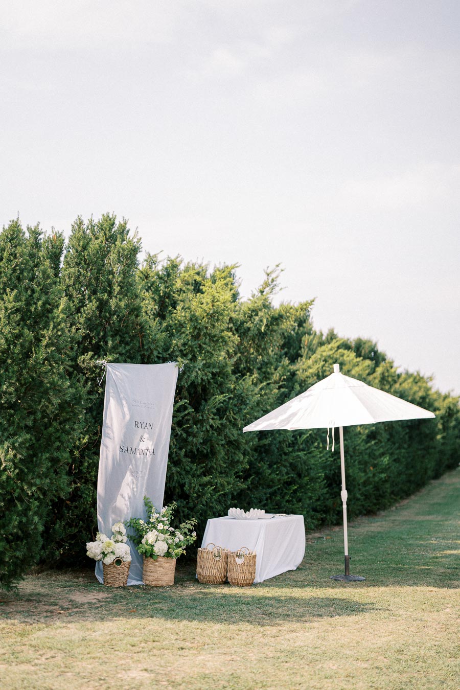 Outdoor wedding setup with elegant floral arrangements, a white parasol, and a simple table draped in white against lush green hedges.