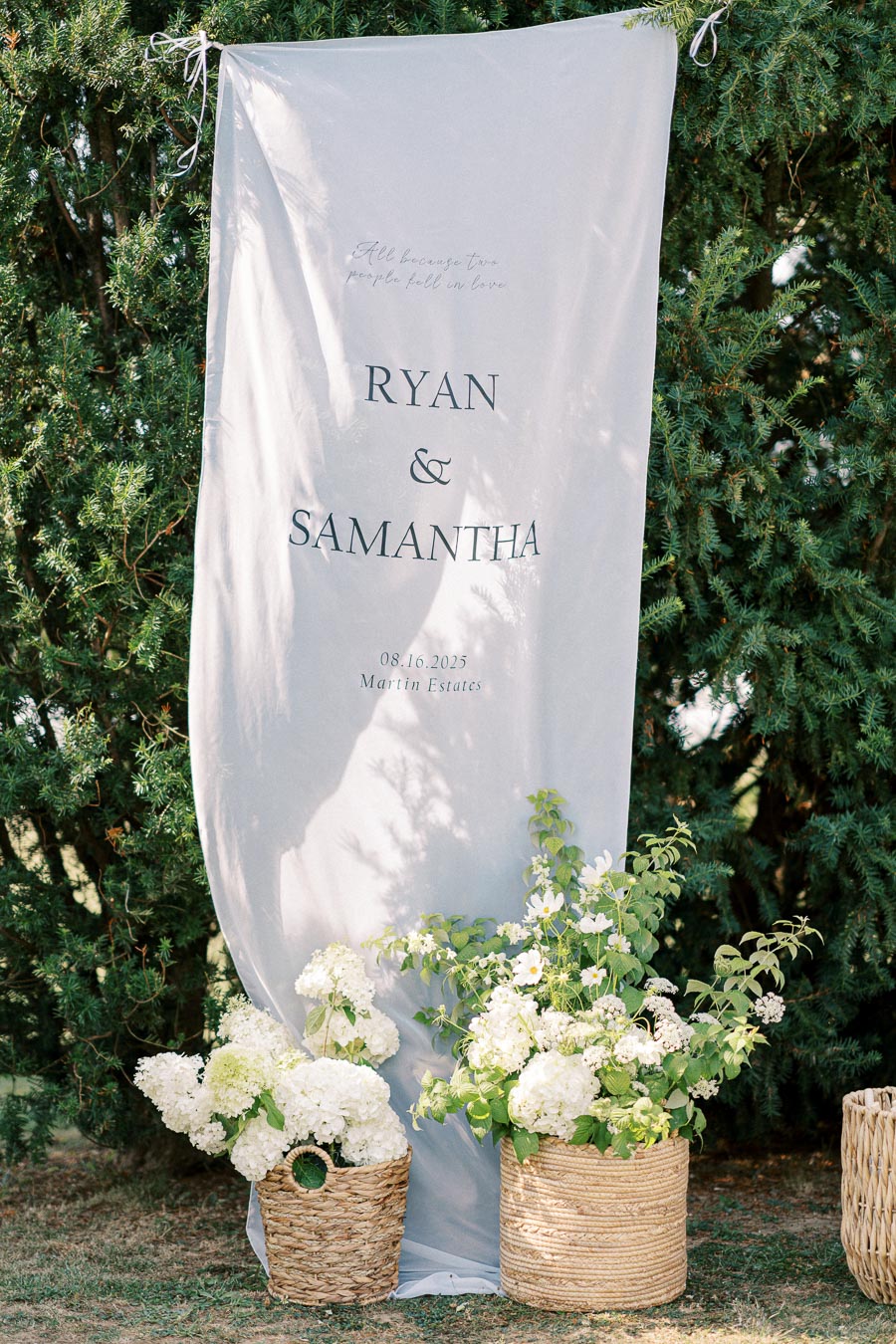 Wedding banner for Ryan and Samantha on August 16, 2025, at Martin Estates, surrounded by lush greenery and white floral arrangements in woven baskets.