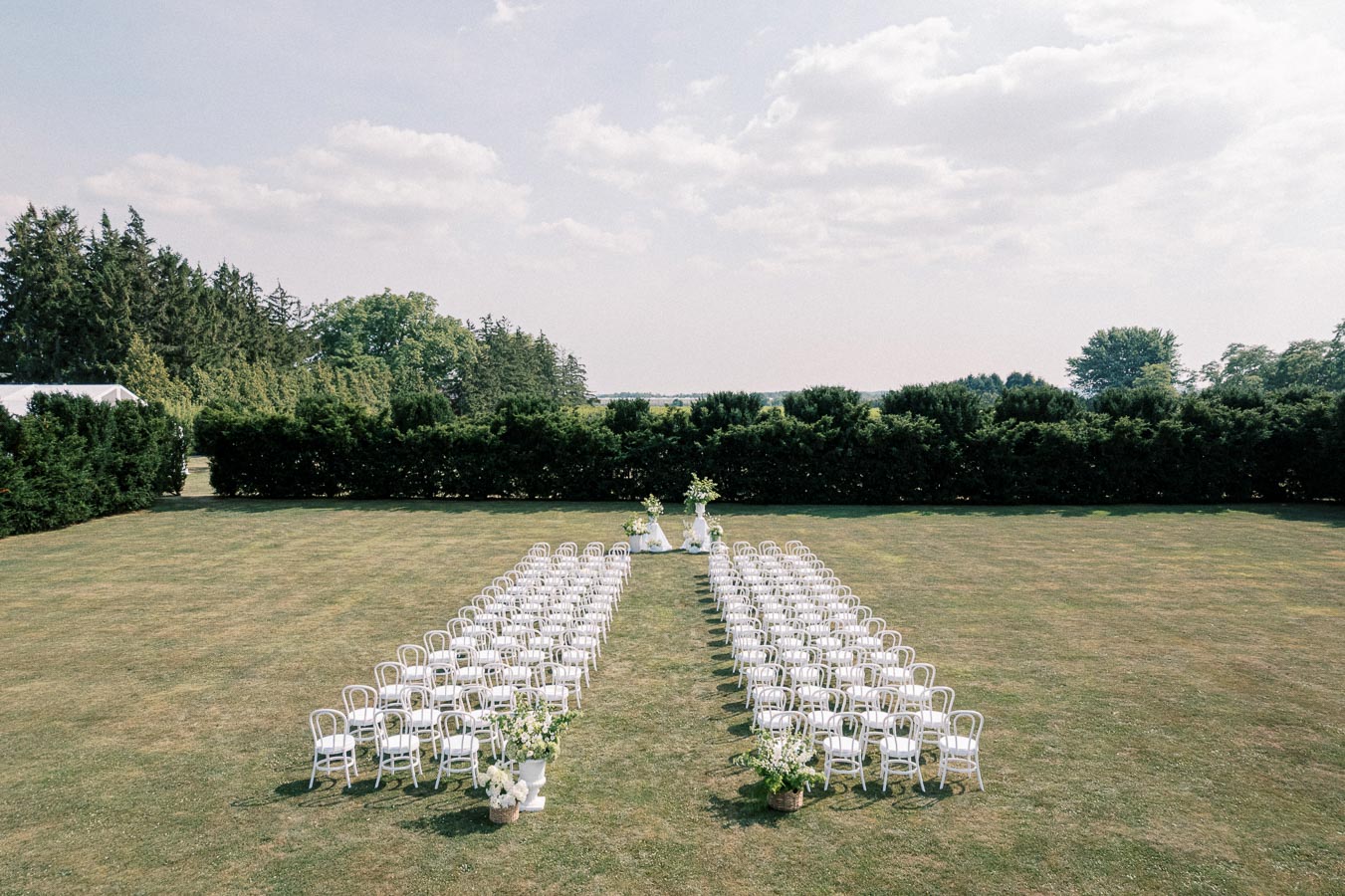 Outdoor wedding ceremony setup with rows of white chairs on a grassy lawn, surrounded by lush greenery and floral arrangements, under a partly cloudy sky.