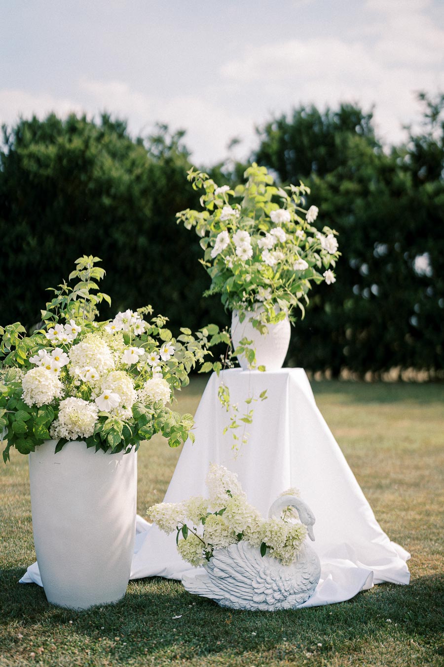 Outdoor wedding decor featuring elegant white flower arrangements in large vases and a swan-shaped planter, set against a lush garden backdrop.