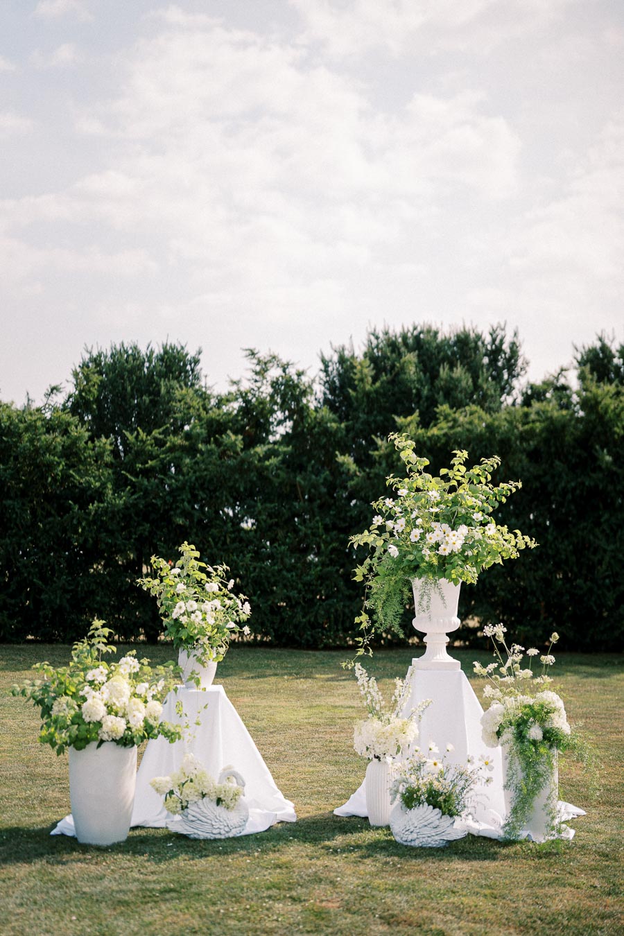 Elegant outdoor garden wedding setup featuring white floral arrangements on pedestals, lush greenery and ceramic swans, against a backdrop of trees and a cloudy sky.