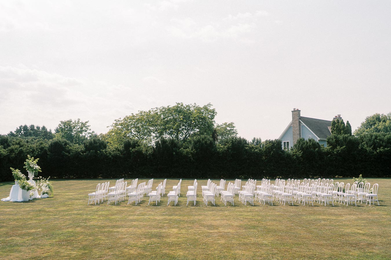 Outdoor wedding ceremony setup in a garden with rows of white chairs facing an altar decorated with greenery and flowers, surrounded by lush trees and a house in the background.