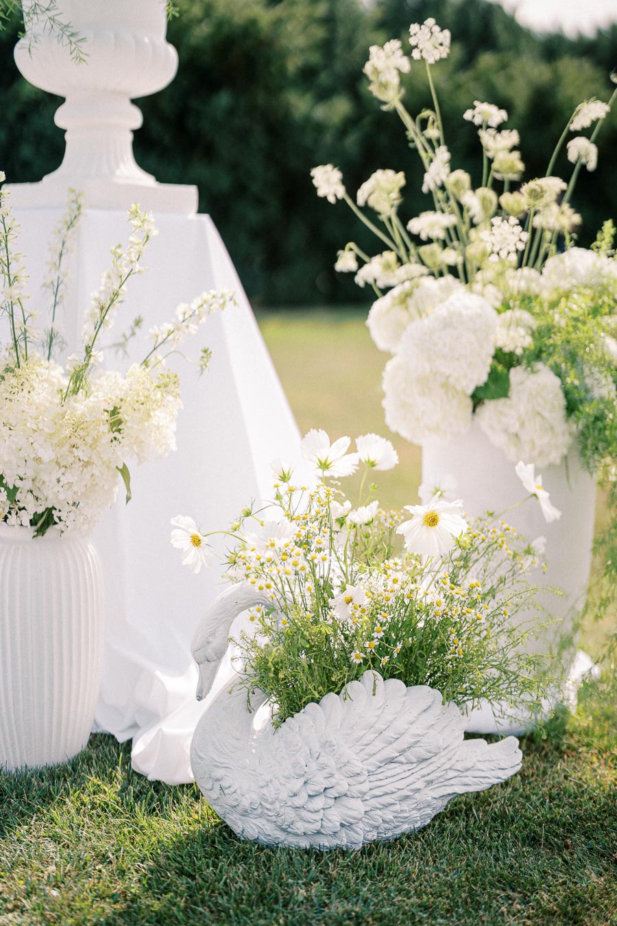 Elegant garden wedding centerpiece with white swan planter, filled with daisies and lush greenery, set on a manicured lawn alongside tall white floral arrangements.