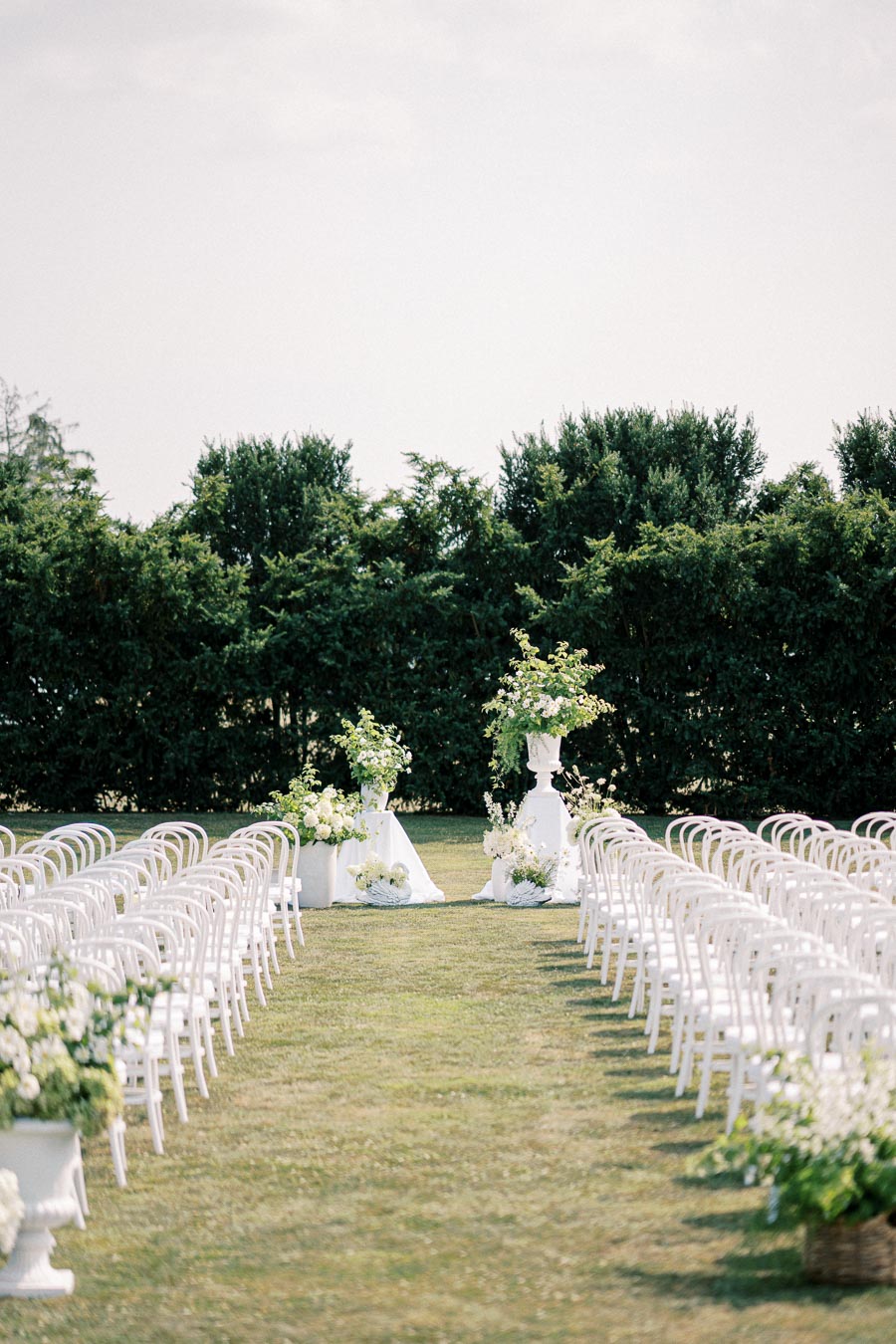 Outdoor wedding ceremony setup with rows of white chairs lining a grassy aisle, bordered by lush greenery and elegant floral arrangements at the altar.