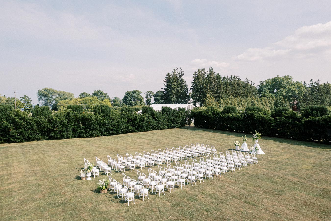Outdoor wedding ceremony setup with rows of white chairs arranged on a grassy lawn, surrounded by lush green trees, under a clear sky.