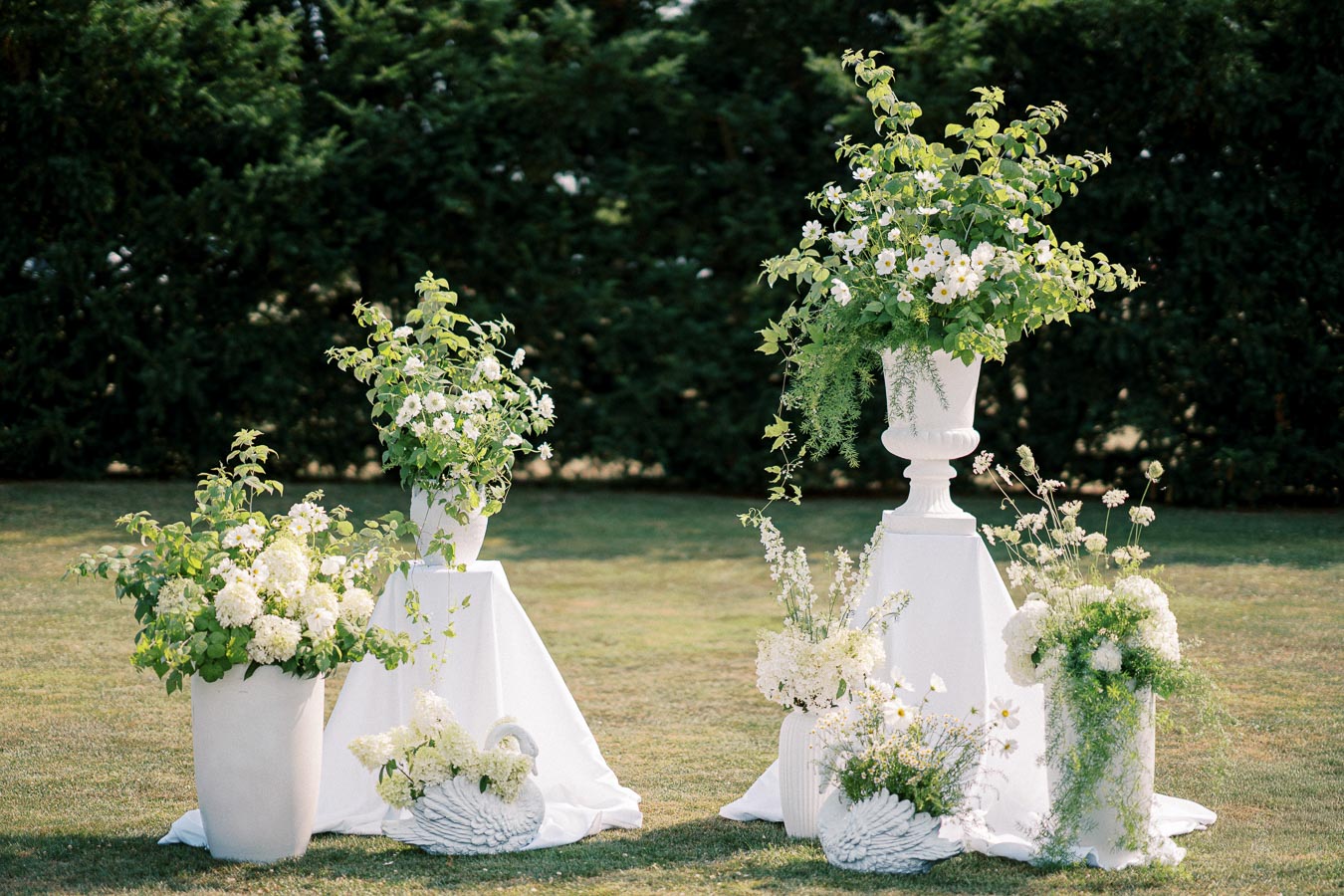 Elegant outdoor wedding floral arrangements featuring white flowers and lush green foliage in various white planters, set on a grassy lawn with a backdrop of dark green shrubs.