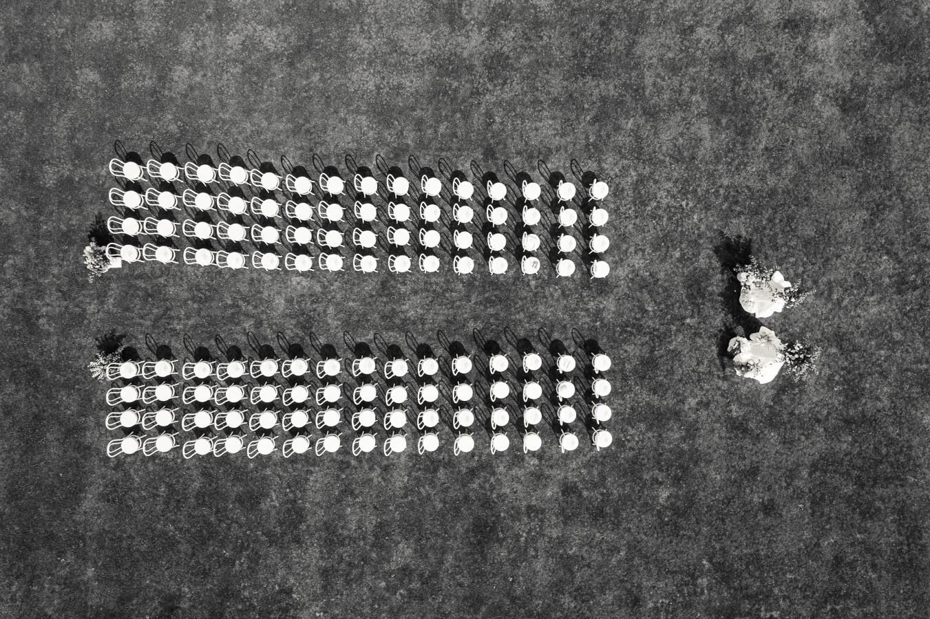 Aerial view of a neatly arranged outdoor wedding ceremony setup with two rows of white chairs on green grass and two brides with bouquets standing to the right, creating a visually striking geometric pattern.
