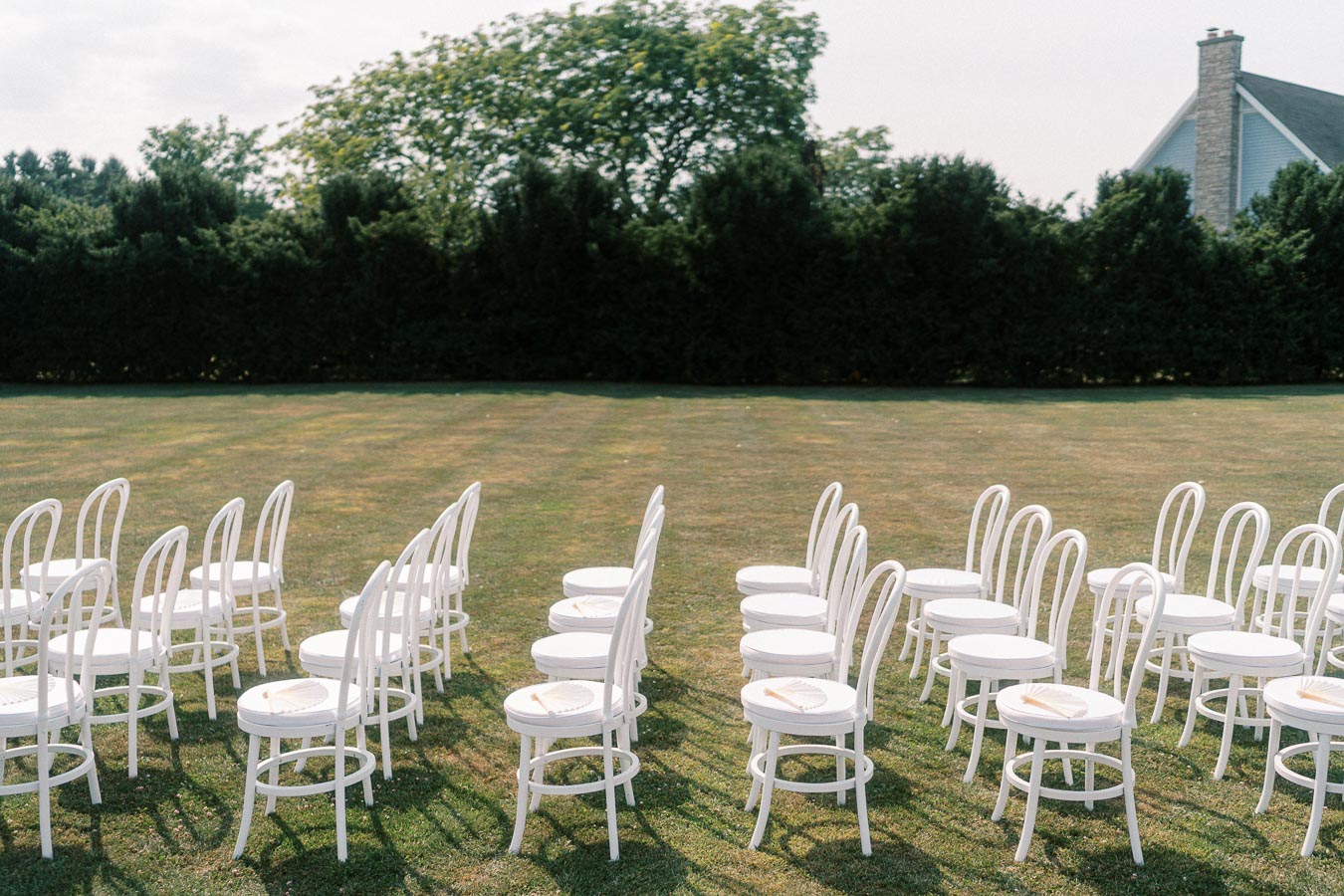Rows of white chairs arranged on a grassy lawn, set up for an outdoor wedding ceremony, with a backdrop of lush green trees and a house in the distance.