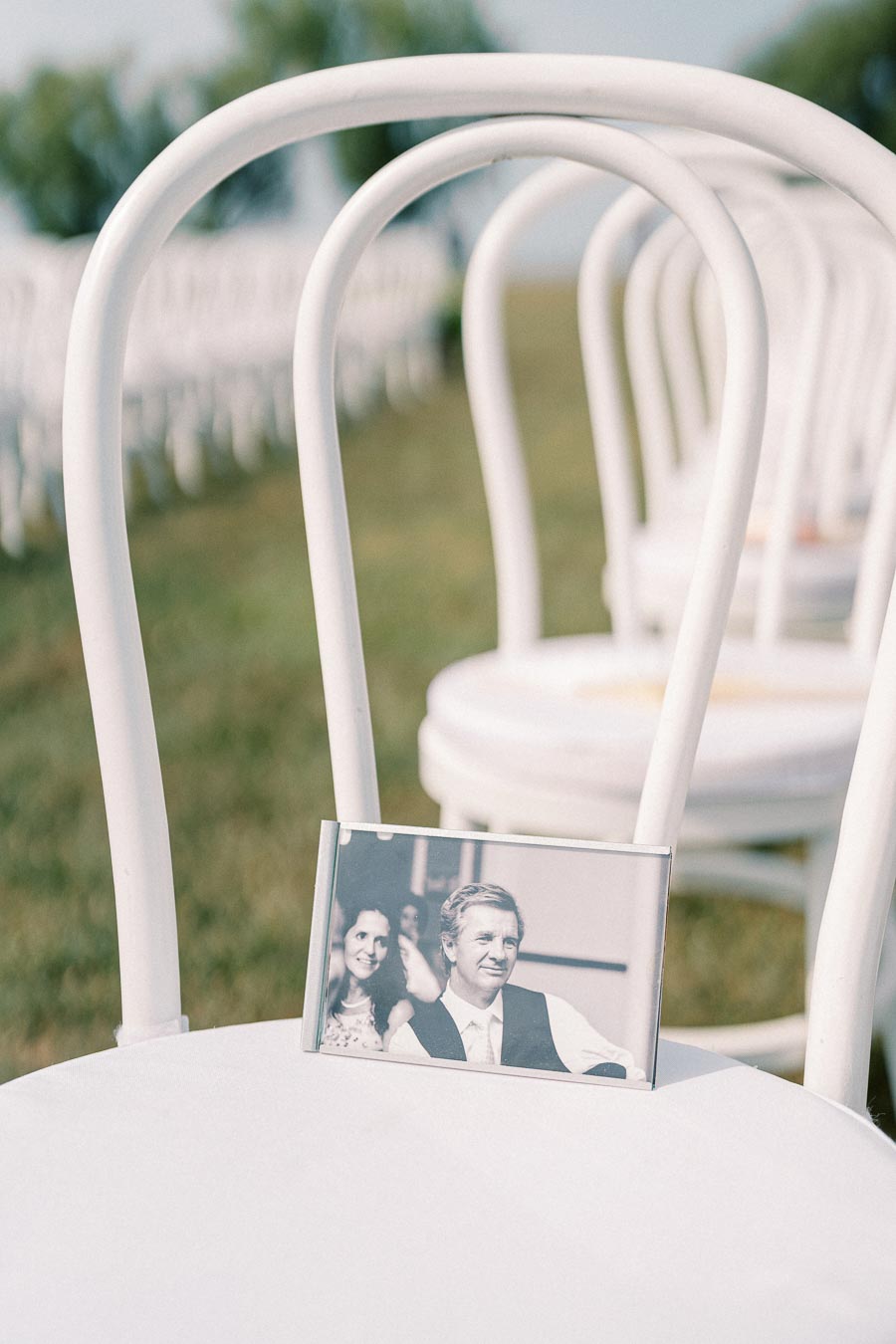 White chair at an outdoor wedding venue with a framed black-and-white photo of a couple placed on the seat, suggesting reserved seating or a memorial tribute.