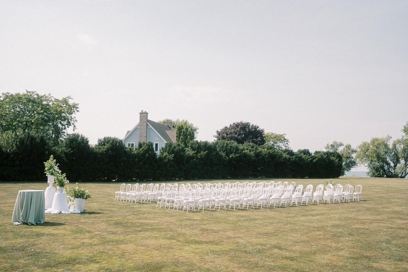 Outdoor wedding ceremony setup with white chairs on a grassy lawn, surrounded by greenery and a quaint house in the background.