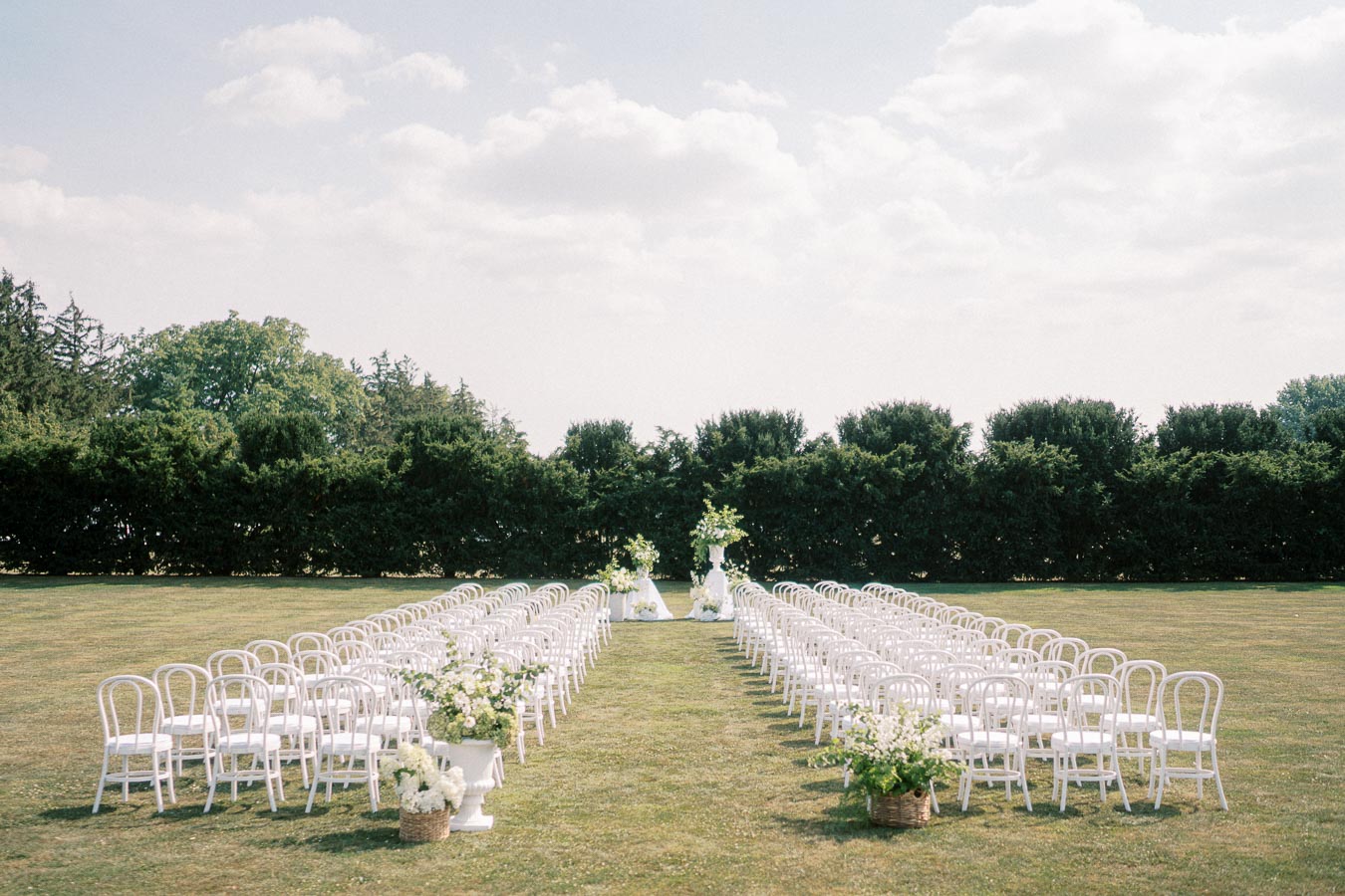 Outdoor wedding ceremony setup with two rows of white chairs and floral decorations on a grassy lawn, surrounded by lush green trees under a partly cloudy sky.