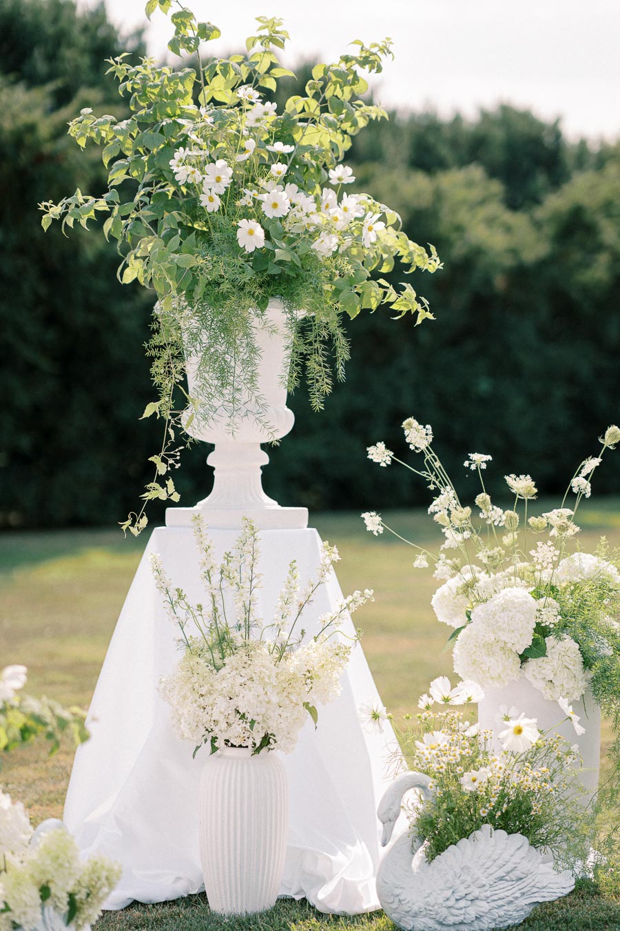 Elegant outdoor flower arrangement featuring white daisies, hydrangeas, and delicate greenery displayed in white vases and swan-shaped planter on a sunny day.