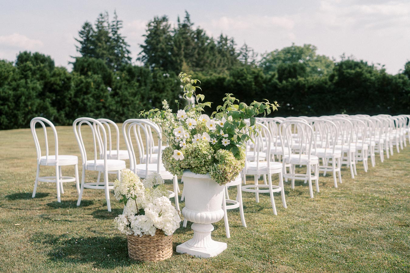 Outdoor wedding ceremony setup with rows of white chairs on a grassy lawn, featuring elegant floral arrangements in baskets and urns with white and green flowers, surrounded by lush greenery.