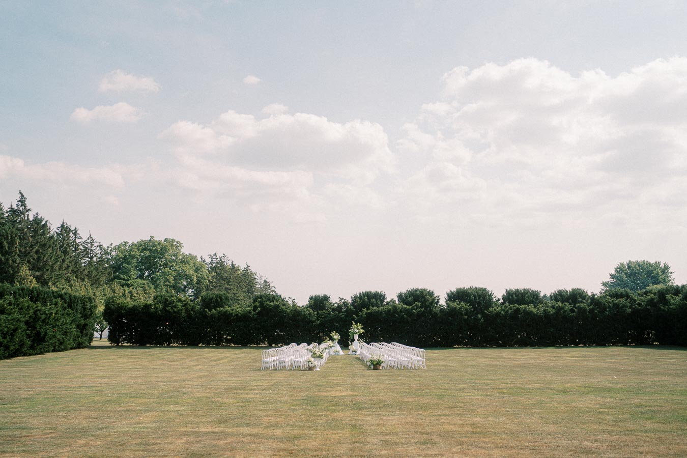 Outdoor wedding ceremony setup on a spacious lawn with rows of white chairs arranged neatly under a partly cloudy sky, surrounded by lush greenery and trees in the background.