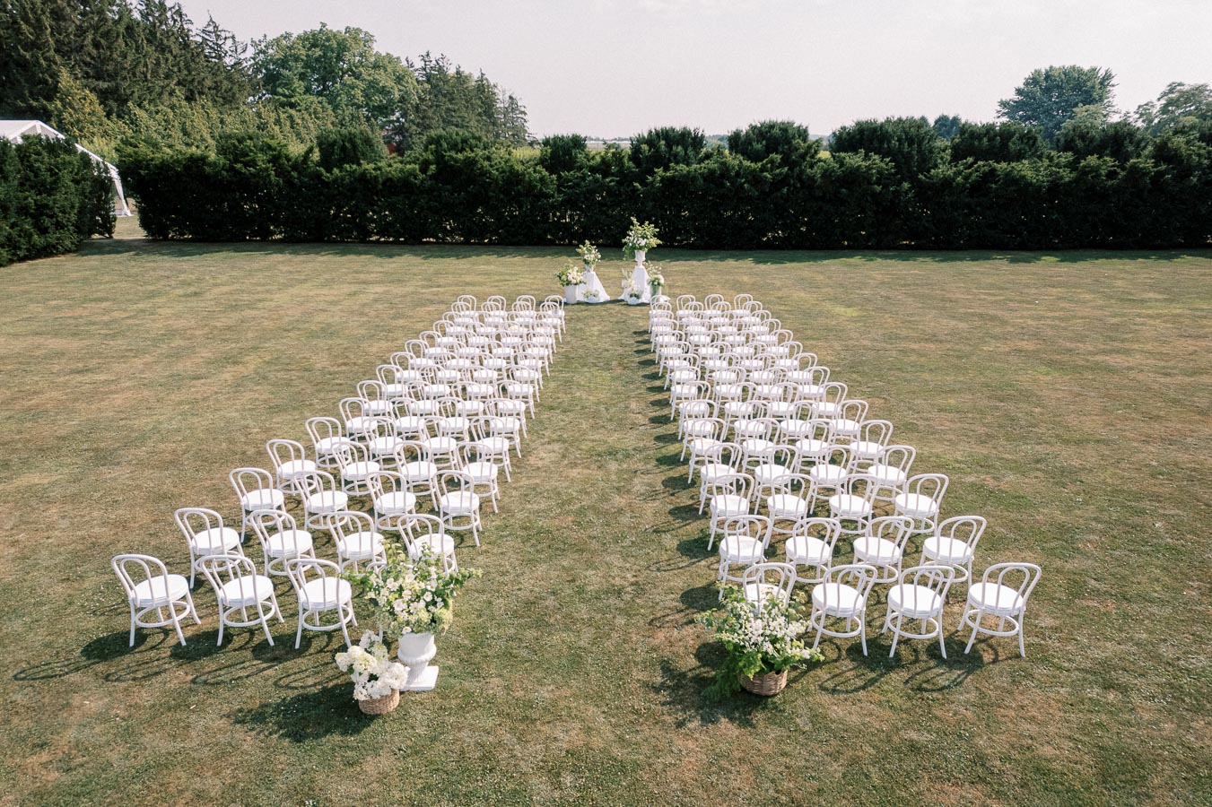 Outdoor wedding ceremony setup with rows of white chairs arranged on a grassy lawn, surrounded by greenery and floral decorations.
