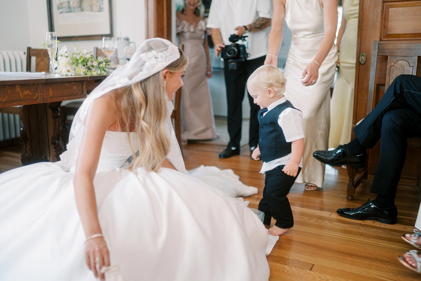 Bride in white dress kneeling to interact with young child in formal outfit during wedding preparation, surrounded by family and friends in an intimate room setting.