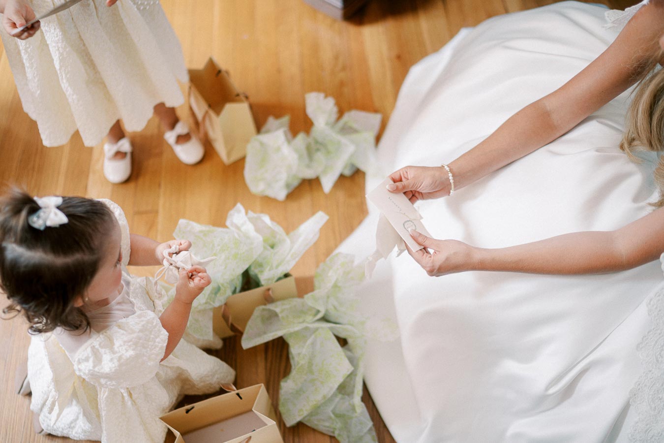 Wedding preparation moment with bride in a white dress and young girl opening gift boxes on a wooden floor.