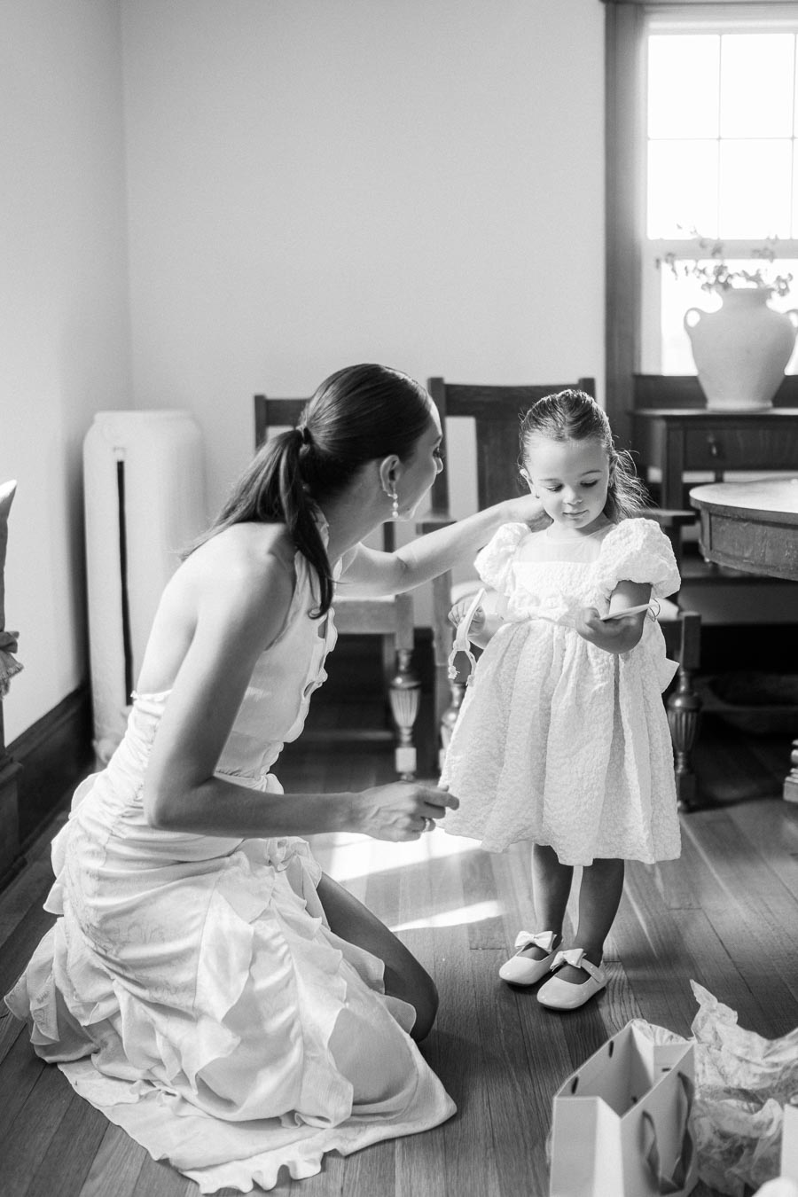 Black and white image of a woman kneeling next to a young girl in a white dress, set in a sunlit room with wooden floors and elegant furnishings, conveying warmth and tenderness.