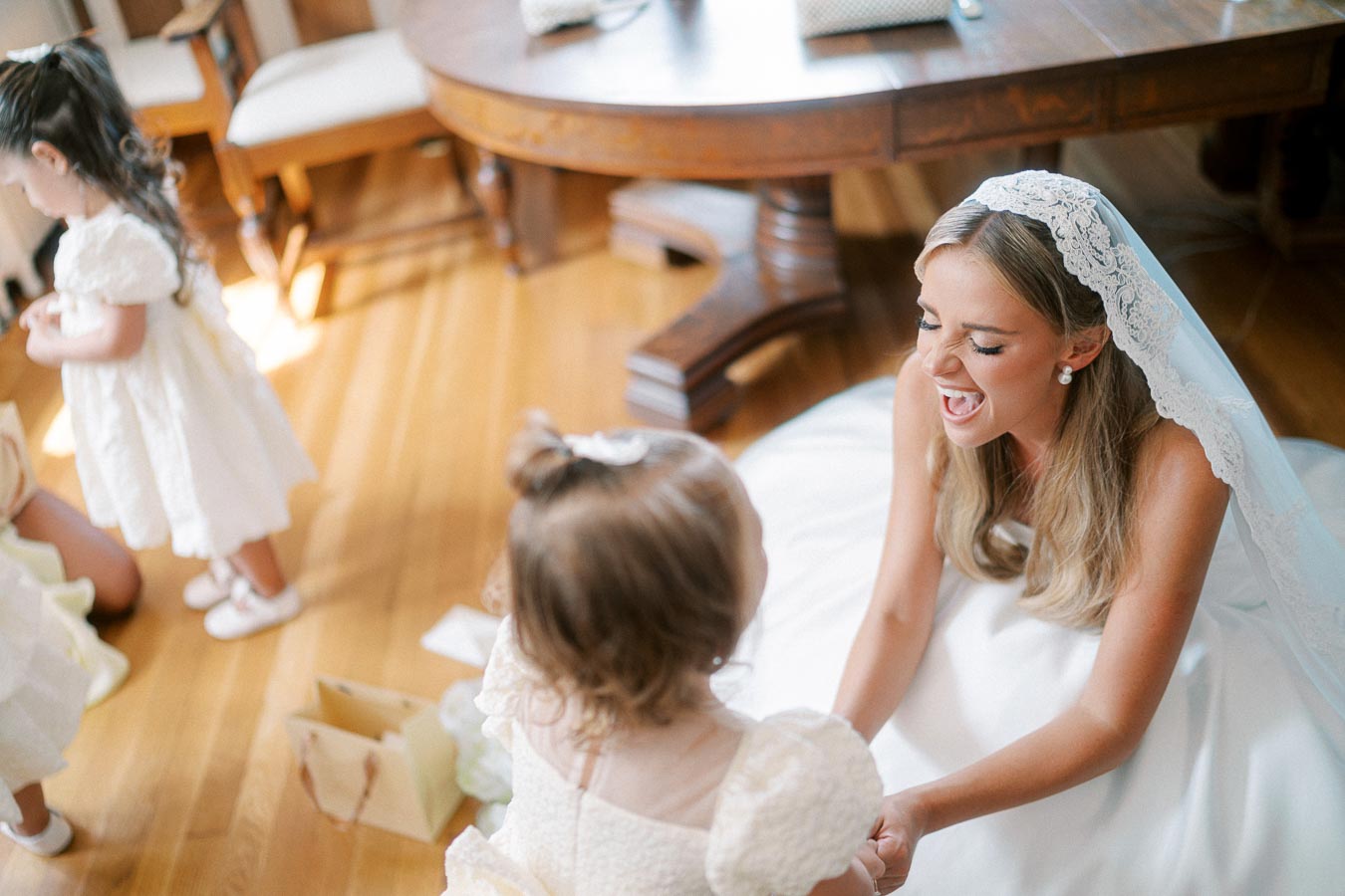 A bride in a white dress and lace veil joyfully interacts with a young flower girl in a bright, elegant room.