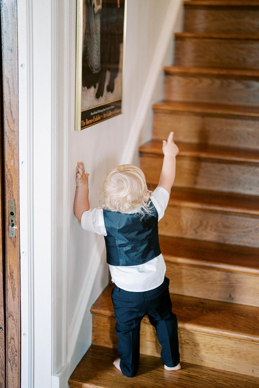 Toddler in a black vest and white shirt climbing wooden stairs, pointing at a framed wall poster.