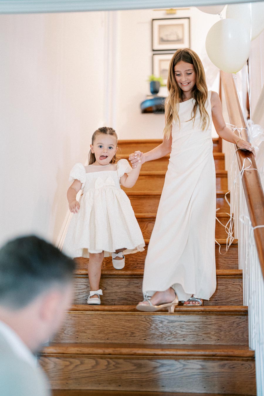 Two young girls in white dresses walking down a wooden staircase, holding hands, with balloons in the background.