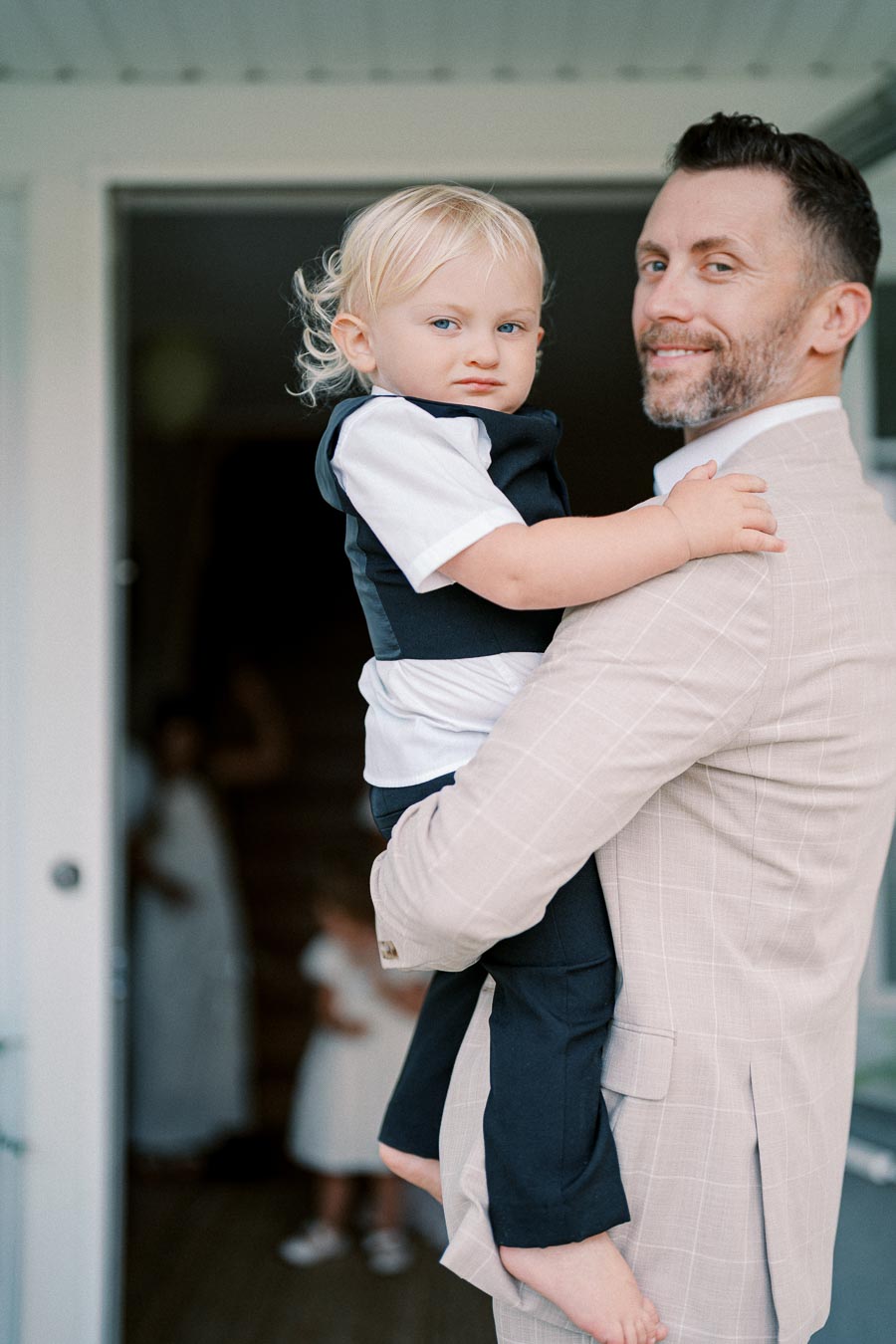 Father holding young child outside a home, both dressed formally, smiling at camera.