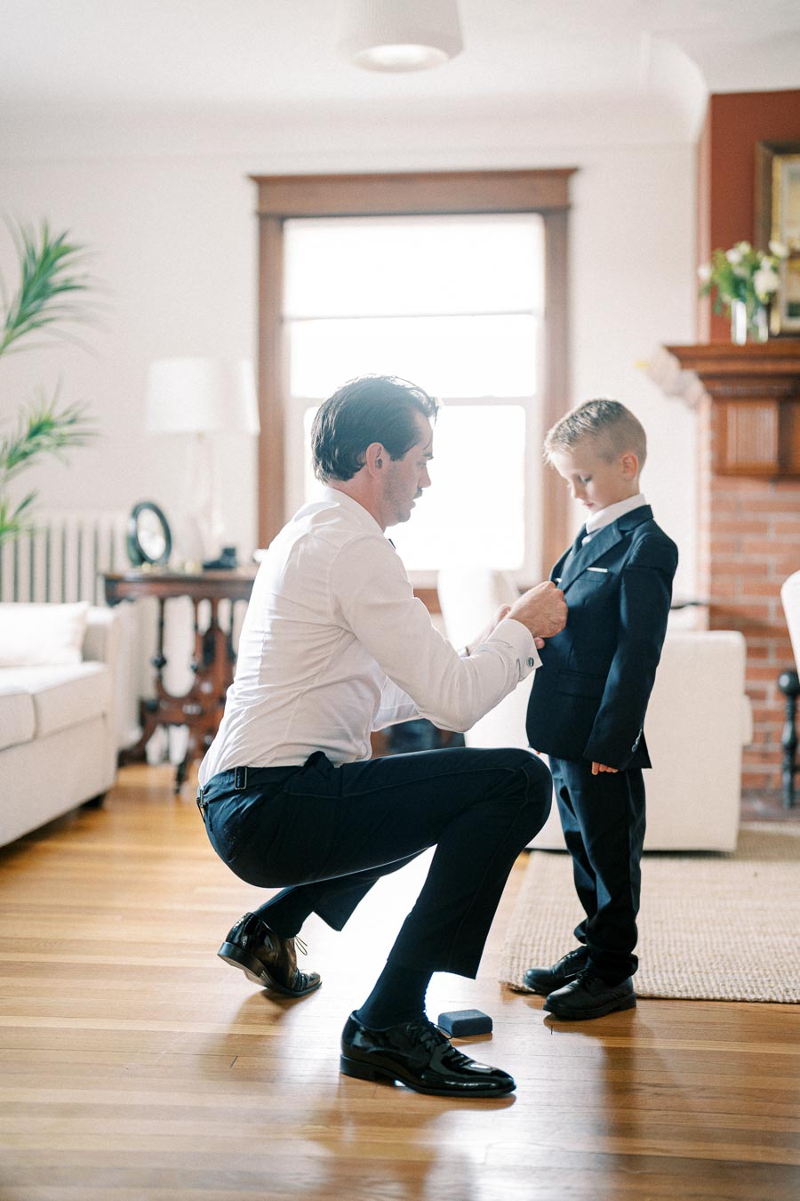 A man kneeling to adjust a young boy's suit jacket in a living room, emphasizing father-son bonding and preparation for a formal event.