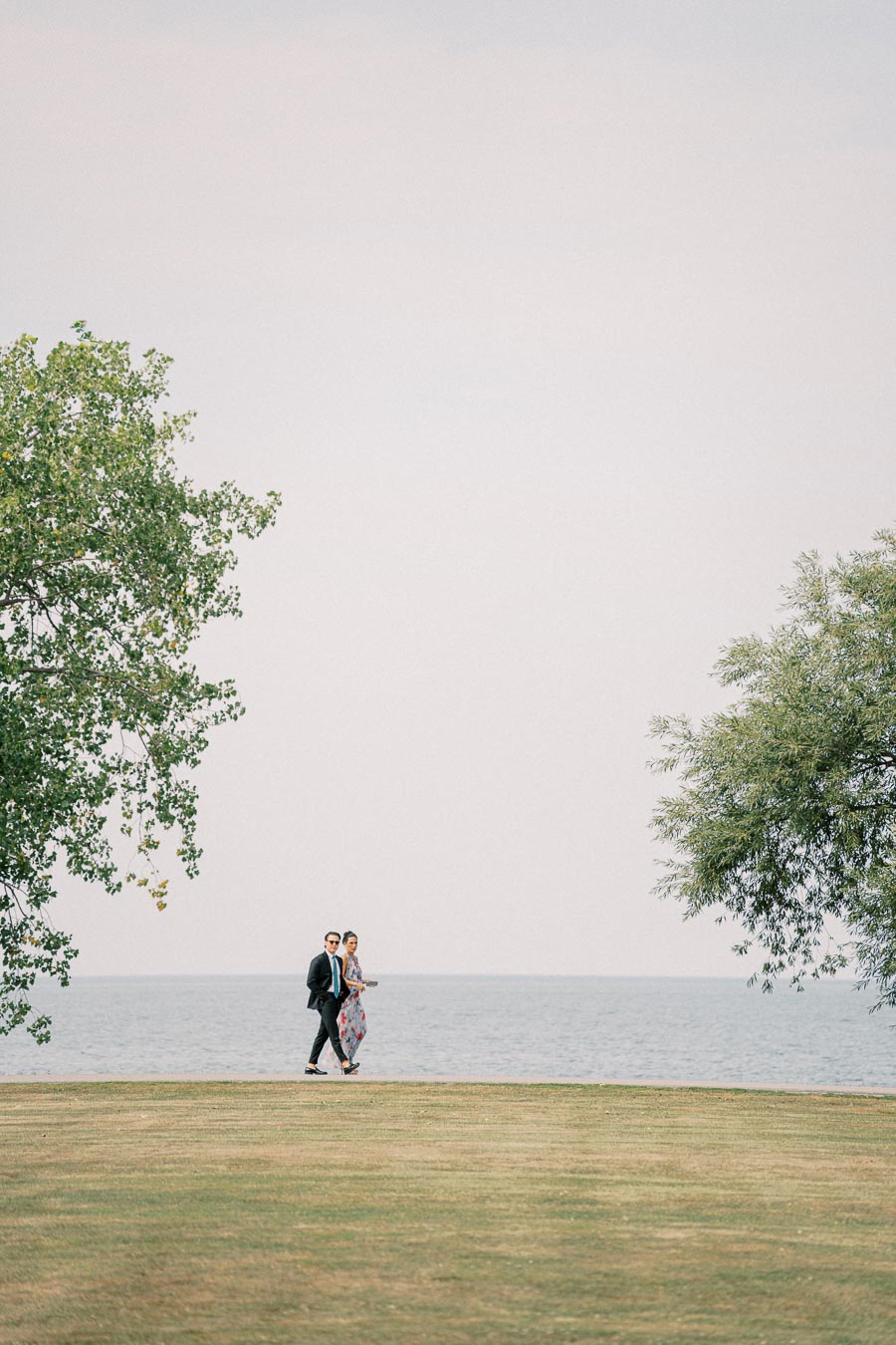 A couple walking on a grassy field between two trees, with a calm sea and clear sky in the background, conveying a peaceful outdoor scene.