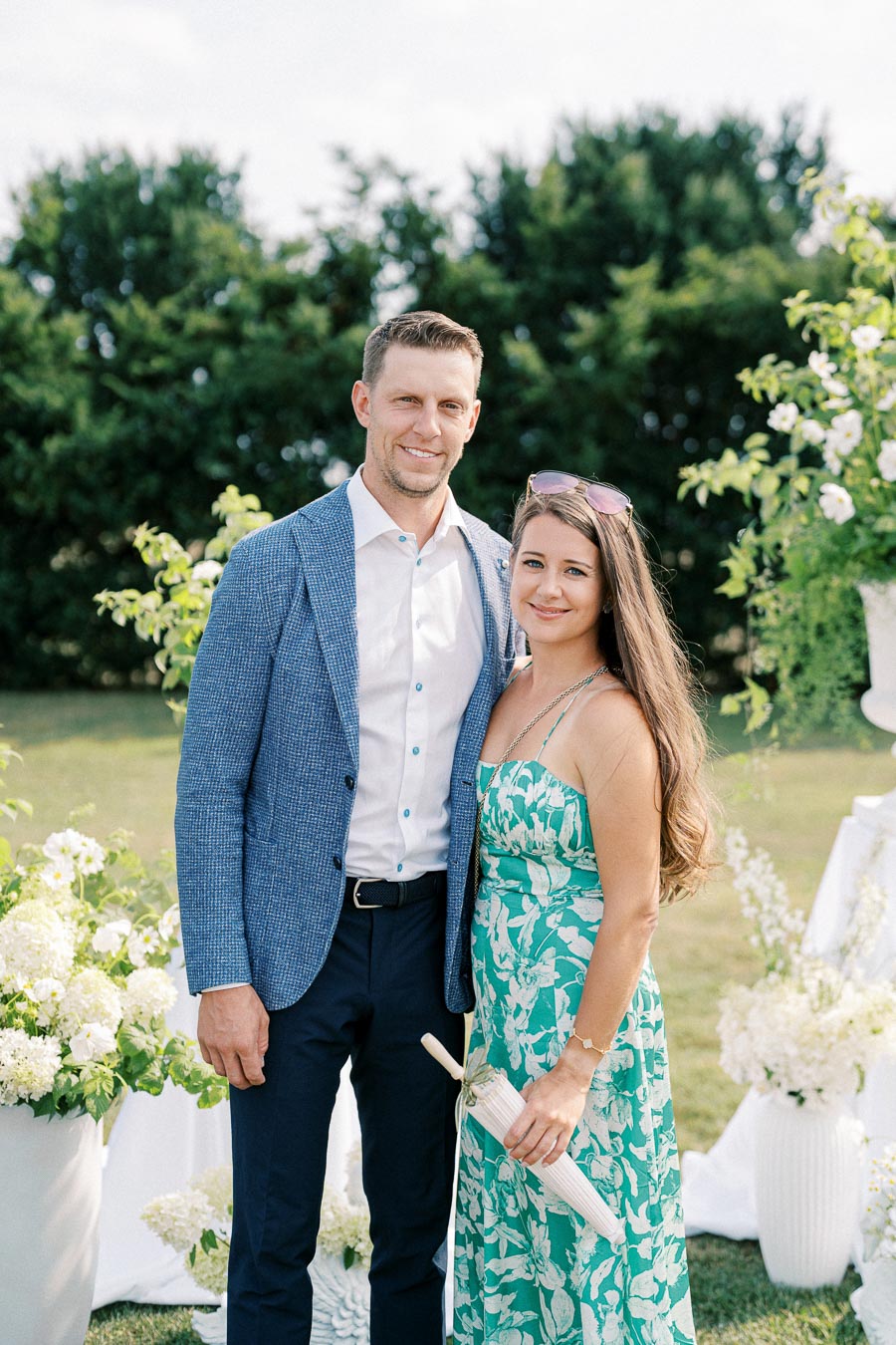 A couple smiling and standing together outdoors in a garden setting, with the woman holding a parasol and vibrant greenery in the background. The man is in a blue blazer, and the woman wears a floral turquoise dress.