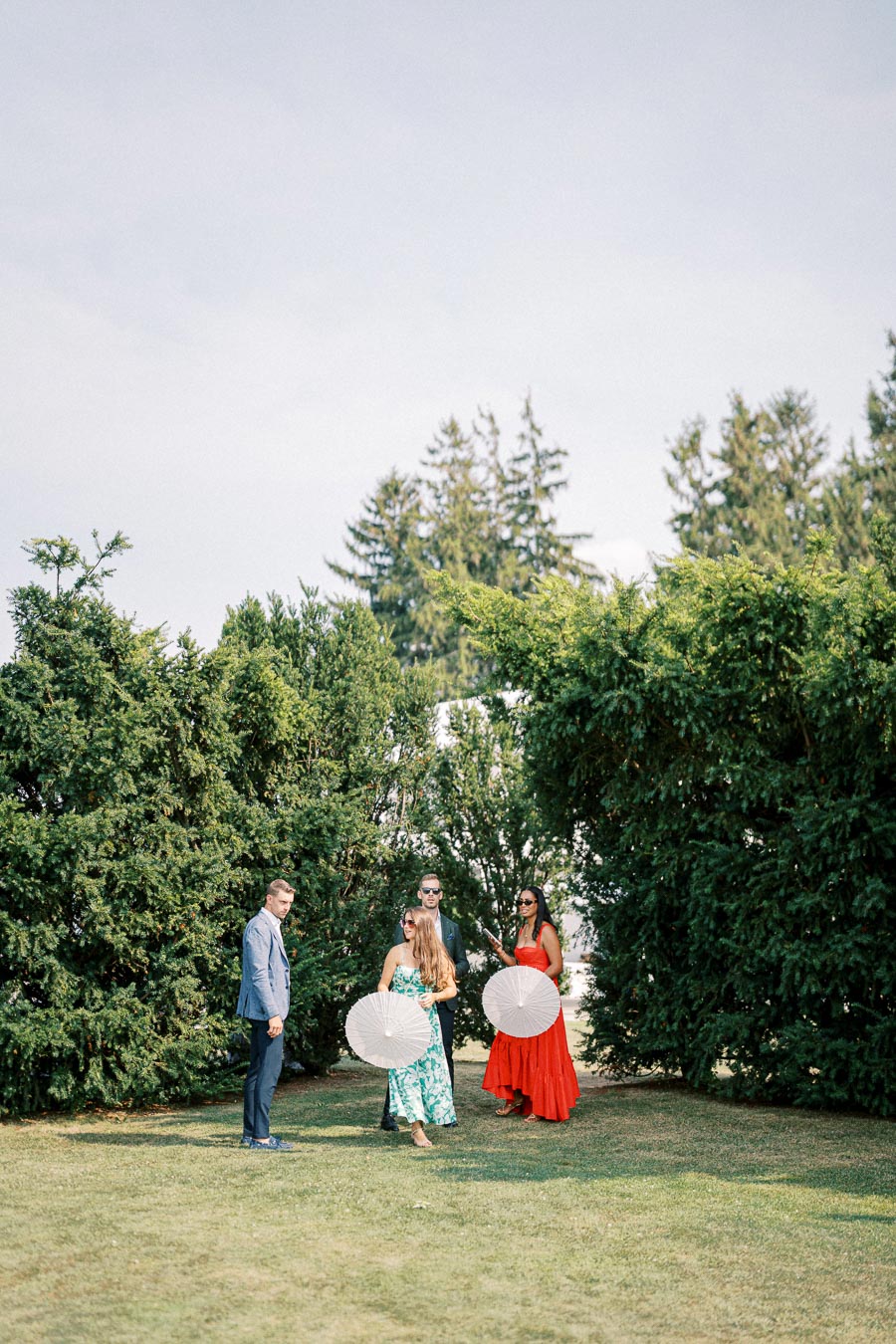 A group of elegantly dressed people standing outdoors on a sunny day, with two individuals holding white parasols, surrounded by lush greenery and tall trees.