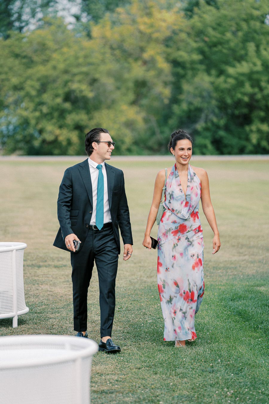 A man in a suit and a woman in a floral dress walk together outdoors on a sunny day, surrounded by lush greenery.