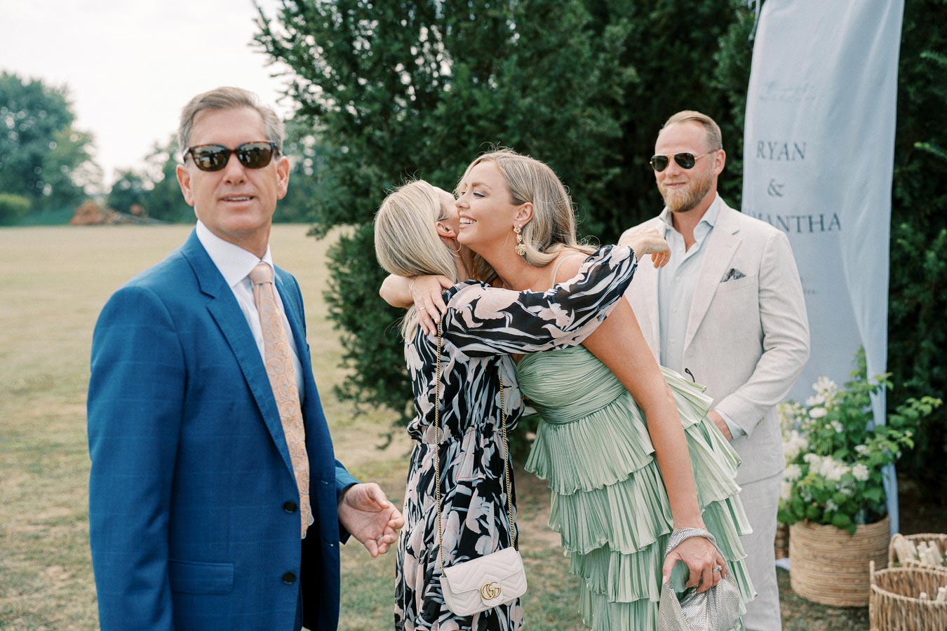 Guests in elegant attire enjoying an outdoor wedding event, with a woman in a green dress hugging another guest, while two men in suits and sunglasses look on, surrounded by greenery and a decorative banner.