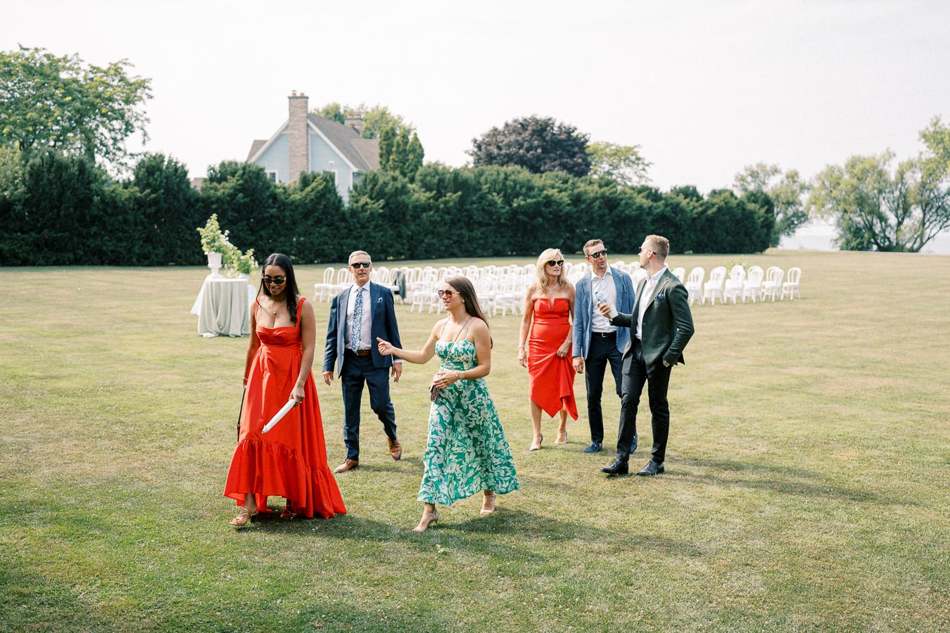 A group of elegantly dressed people walking across a grassy field with a house and rows of chairs in the background, suggesting a gathering or outdoor event on a sunny day.