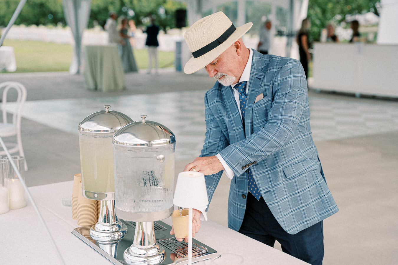 An elderly man in a blue checkered suit and straw hat pours a drink from a beverage dispenser at an outdoor event, featuring a beautifully decorated table setup with cups and drink containers.