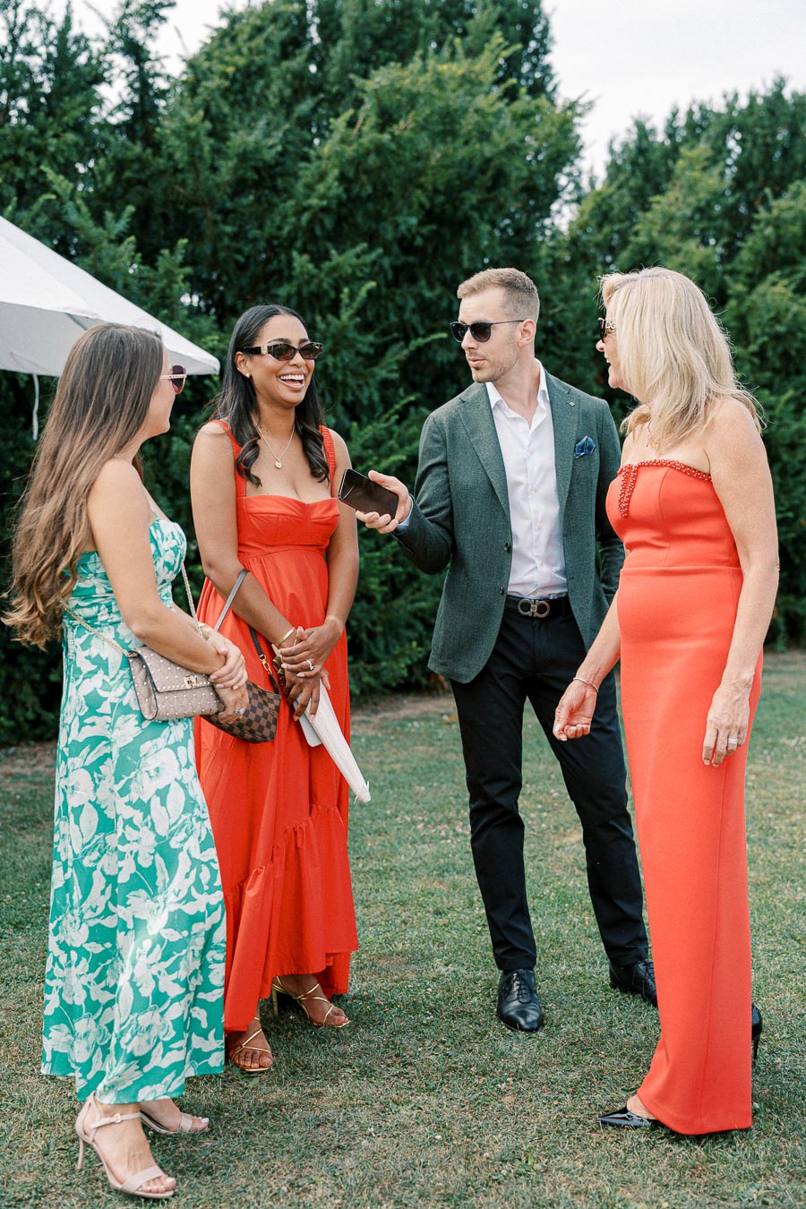 Group of elegantly dressed people socializing outdoors, three women in colorful dresses and one man in a suit, enjoying a garden gathering event.