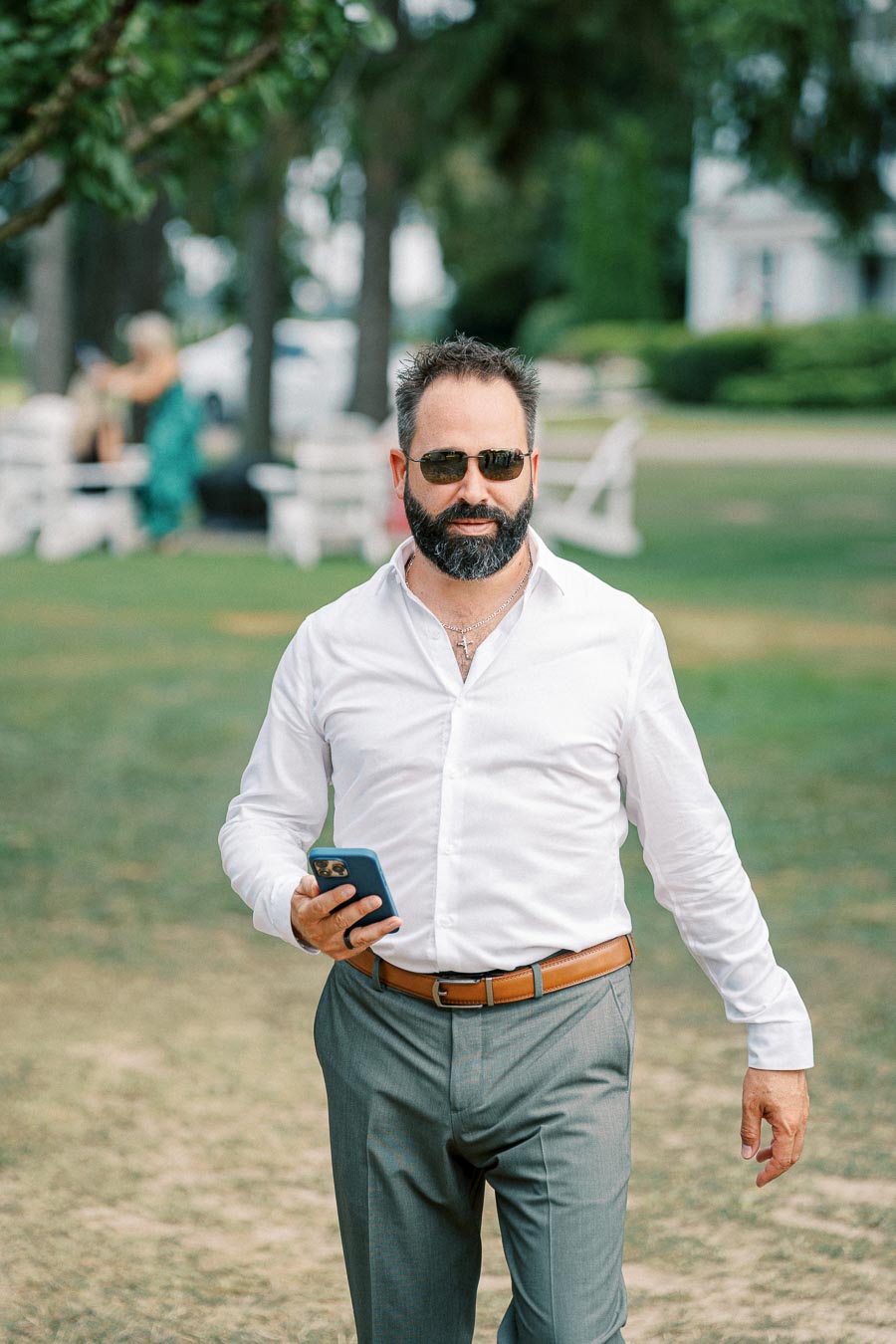 Confident man in sunglasses walking on a grassy field, wearing a white shirt and holding a smartphone