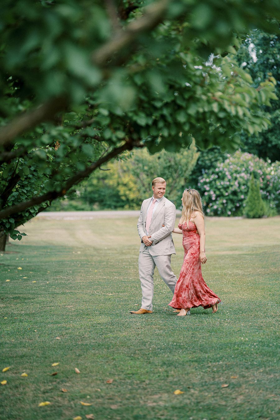 A couple walking through a lush garden, with a man in a light suit and a woman in a flowing red dress, capturing a serene outdoor moment.