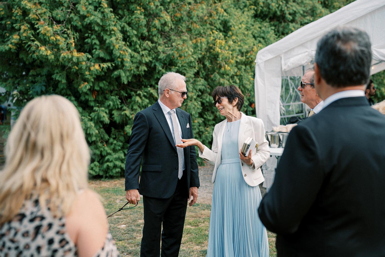 A group of elegantly dressed people socializing outdoors at an event, with a man in a suit and sunglasses conversing with a woman in a light blue dress and white blazer, surrounded by greenery and a white tent canopy in the background.