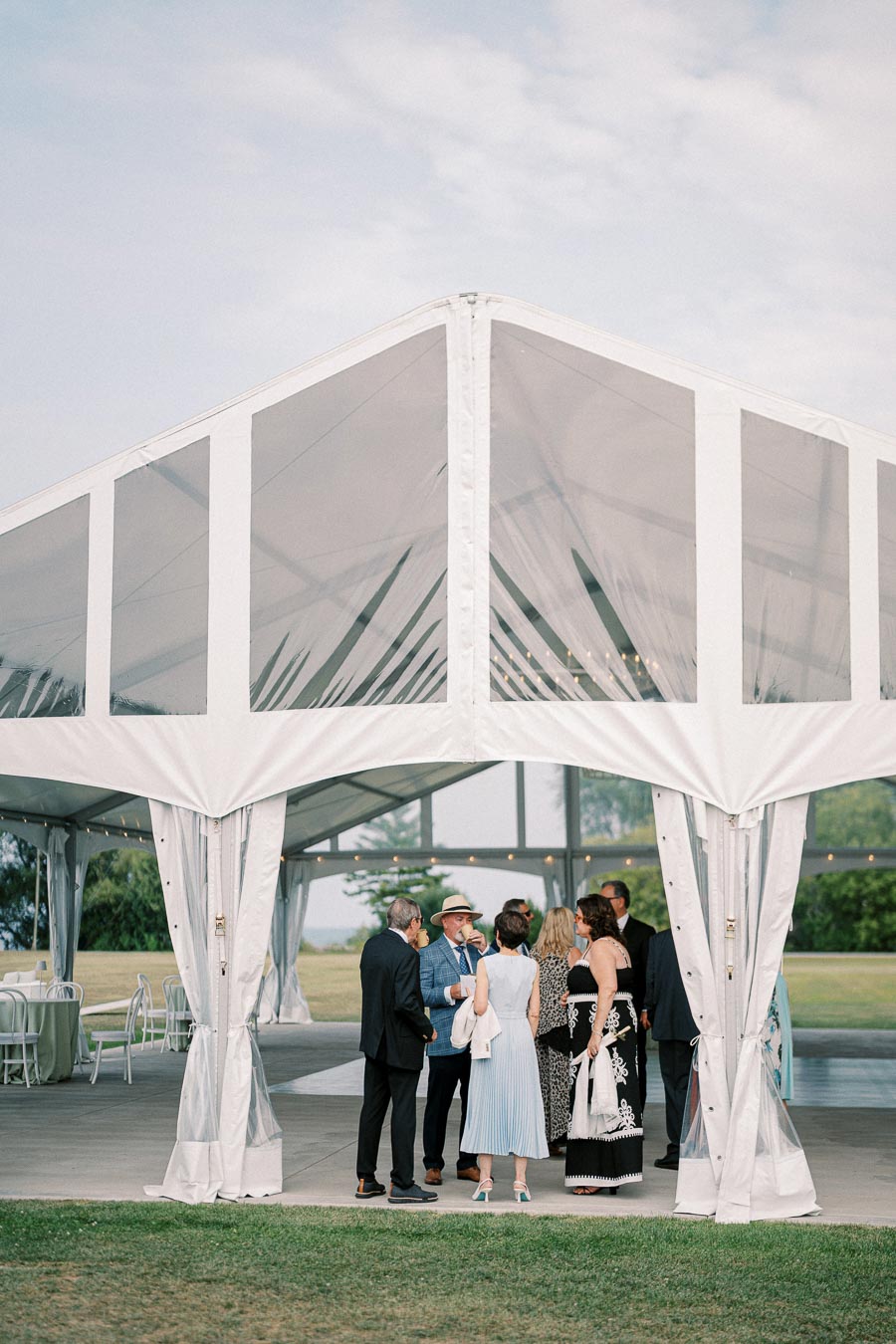 Outdoor event gathering under a white marquee tent, with elegantly dressed guests socializing on a sunny day.
