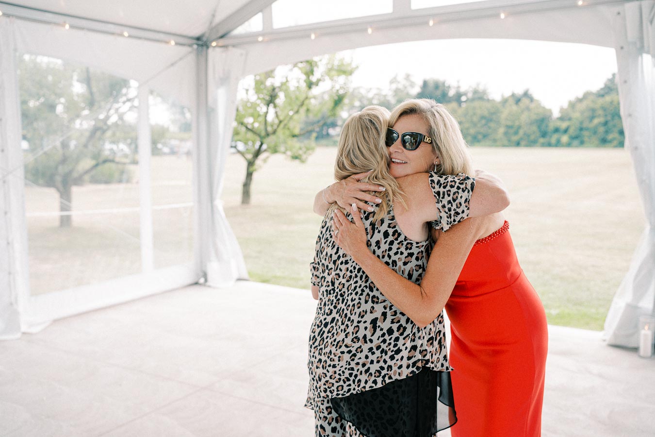 Two women embracing under a white tent in a grassy outdoor setting, with one wearing a red dress and the other in a leopard print top.