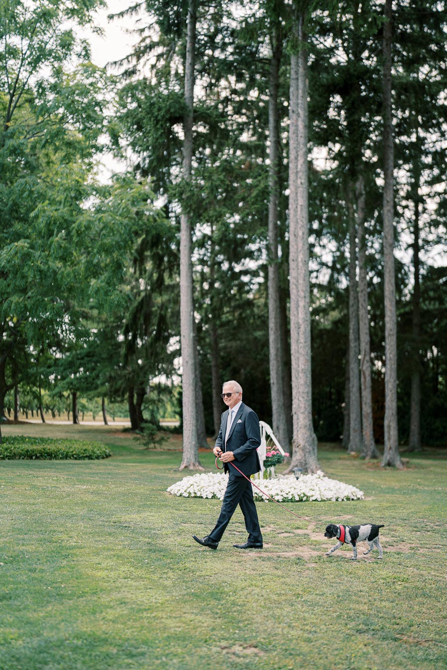 A man in a suit walks a small dog on a leash through a lush park, surrounded by tall trees and a flower bed, creating a peaceful outdoor scene.