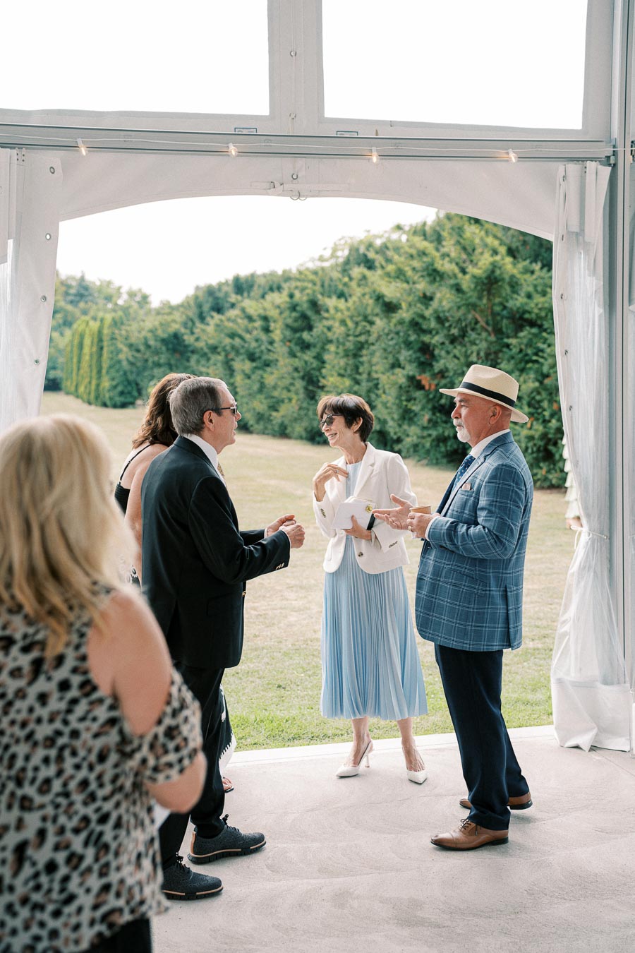 Group of people engaged in conversation at an outdoor event, with lush greenery in the background, under a white event tent.
