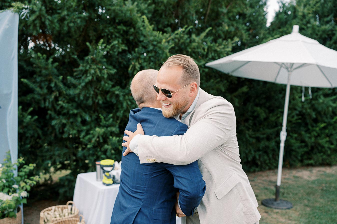 Two men embracing warmly at an outdoor event, with one wearing sunglasses and a light suit, and the other in a blue suit, surrounded by greenery and a white umbrella.