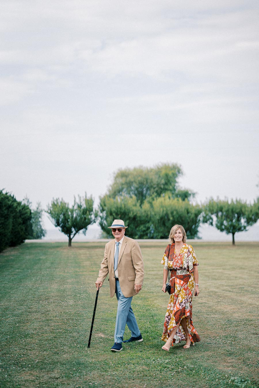 Elderly couple in stylish attire walking in a grassy park on a sunny day, with trees in the background.